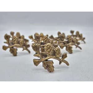 May include: Set of brass-toned napkin rings. Each ring features a detailed design of a branch with leaves and pinecones. The rings are arranged on a white surface, showcasing their decorative nature.