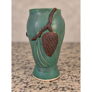 May include: A teal ceramic vase featuring a raised, textured brown pinecone and pine needle relief design on the front. The vase has a smooth, matte finish, a flared rim, and a tapered base, showcasing a distinct botanical motif in relief.