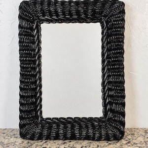 May include: A rectangular wall mirror featuring a glossy black woven wicker frame. The frame is constructed with a dense, braided pattern that surrounds the central reflective glass surface, creating a uniform and textured border around the mirror.