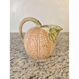 May include: A ceramic pitcher shaped like a cantaloupe melon with a textured, light orange, netted rind surface. The handle is designed to resemble a green vine, and a green leaf is sculpted near the spout, adding a naturalistic detail.