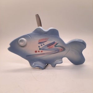 May include: A light blue ceramic fish-shaped spoon rest with a white eye and abstract pink, red, and blue painted details. A silver spoon is inserted into the top of the fish. The spoon rest is a decorative kitchen accessory.