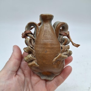 May include: A small, brown ceramic vase with three handles and a leather strap. The vase has a rustic, handmade look.