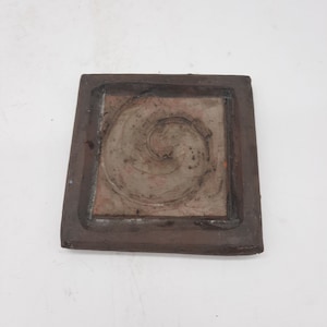 May include: A square ceramic tile with a raised, brown and white swirl design in the center. The tile is framed by a dark brown border.