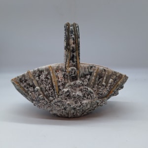 May include: A ceramic basket with a textured, speckled surface in shades of gray, white, and brown. The basket has a handle and a fan-shaped body with a decorative design. The rim features gold accents.