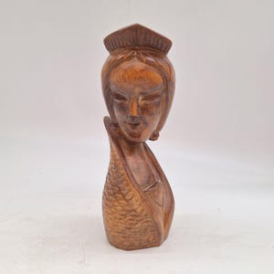 May include: A carved wooden sculpture of a woman's head and upper body. The sculpture is a warm brown color, with detailed carving of the face, hair, and clothing. The woman's eyes are closed, and she has a serene expression. The sculpture has a textured, patterned design on the lower portion.