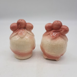 May include: A pair of vintage ceramic salt and pepper shakers. The shakers are shaped like bundles with a light beige base and pink accents. Each shaker has a cluster of pink spheres at the top with holes for dispensing.