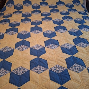 May include: A handmade quilt with a geometric pattern of blue and yellow shapes. The blue shapes are filled with a floral pattern. The quilt is likely made of cotton and has a quilted texture.