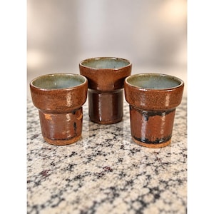 May include: Three ceramic cups with a glossy, dark brown exterior glaze and a contrasting light blue interior glaze. Each cup features a distinct flared rim and a cylindrical base, showcasing a textured, earthy finish with subtle dark markings.