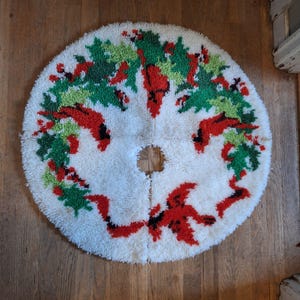 May include: A round, off-white Christmas tree skirt with a central hole. The skirt features a design of red cardinals, green holly leaves, and red berries arranged in a circular pattern. The skirt is made of a textured, looped material.