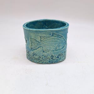 Puede incluir: Un vaso de cerámica turquesa con una superficie texturizada y un diseño de pez. El pez está en relieve en el lateral del vaso. La parte inferior del vaso tiene un diseño texturizado en forma de ola.