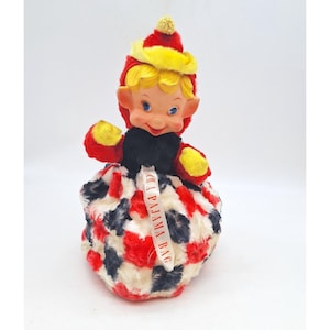 Puede incluir: Una muñeca vintage para bolsa de pijama con una cara sonriente, cabello rubio y ojos azules. La muñeca lleva un sombrero rojo y amarillo y una parte superior negra. La falda tiene un estampado de piel roja, blanca y negra. Una cinta dice "A Pajama Bag."