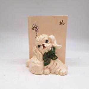 May include: A cream-colored ceramic dog figurine with a green bow, set against a backdrop resembling a book. The dog has black eyes and a pink nose. Floral and insect designs are painted on the book-like background.