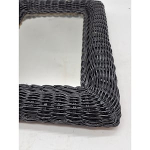 May include: A square mirror with a black woven wicker frame. The frame has a textured, braided appearance. The mirror's reflective surface is visible within the frame. The overall design is simple and rustic.