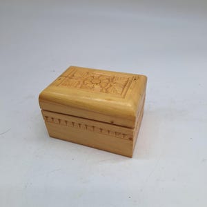 May include: A small, rectangular wooden box with a carved floral design on the lid. The box is light brown and has a simple, elegant design. The edges of the box are decorated with small, triangular carvings. A decorative storage box.