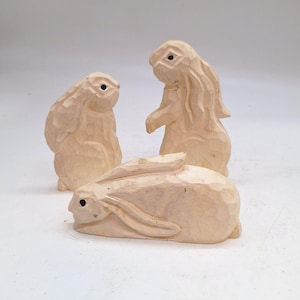 May include: Three wooden rabbit figurines. One rabbit is lying down, while the other two are standing. The rabbits are carved from light-colored wood and have dark eyes. The figurines are set against a white background.