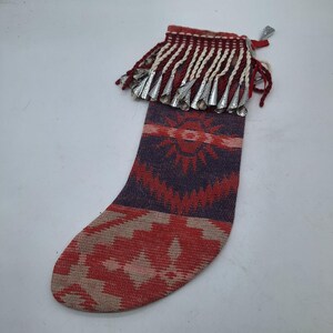 Puede incluir: Un calcetín navideño tejido con un diseño del suroeste. El calcetín presenta un patrón geométrico rojo, morado y beige. La parte superior tiene una franja de borlas blancas y rojas con campanas plateadas. Se adjunta un pequeño lazo plateado para colgar.