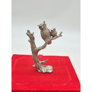 May include: A silver-colored owl figurine with a smaller owl perched on a branch. The figurine is set on a red surface. The owls have detailed feather textures and are facing to the left. The branch is also silver and has a textured appearance.