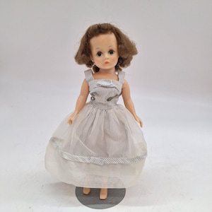 May include: A vintage doll with brown hair and a light blue and white striped dress. The doll has a white sheer skirt and is standing on a gray metal stand. The doll's dress is secured with safety pins.