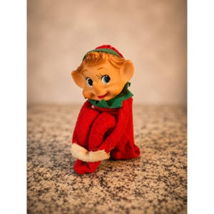 May include: A small elf figurine with a tan face, blue eyes, and a pointed nose. The elf wears a red felt outfit with a green collar and a matching red hat with a green trim. The figure is posed in a seated position with knees pulled up.