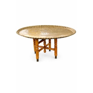 May include: A round, antique brass tray table. The tray has a decorative, scalloped edge and rests on a wooden base with four legs. The legs are light brown with brass-colored feet. The table is a vintage piece.