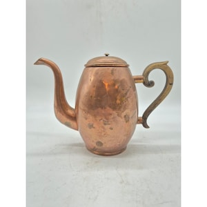 May include: A copper teapot with a curved spout and a lid. The handle is a light brown color. The teapot has a rounded body and sits on a white surface. The teapot is likely vintage or antique.