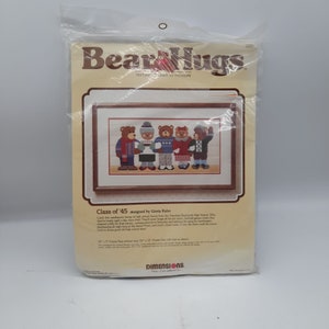 May include: A cross-stitch pattern featuring five teddy bears wearing winter clothing. The pattern is titled "Bear Hugs" and "Class of '45" designed by Gloria Kahn. The pattern is from Dimensions, a brand of needlework kits.
