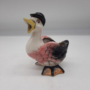 Puede incluir: Figura de pato de cerámica con sombrero negro, pico abierto y detalles en rosa y negro. El pato tiene cuello y pecho blancos, pico y patas amarillas. Un artículo decorativo.