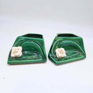 May include: Two green ceramic planters with a glossy finish. Each planter features a white flower accent on the front. The planters have a unique, angled design, perfect for small plants or decorative use.
