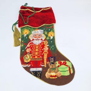 May include: A Christmas stocking with a red velvet cuff and gold tassel. The stocking features a detailed needlepoint design of a nutcracker, a teddy bear, and a wrapped gift. The background is green with gold accents.