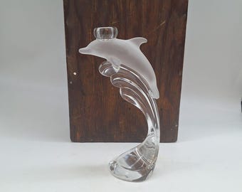 May include: A clear glass dolphin ornament featuring a frosted dolphin and a clear, curved base. The dolphin is depicted leaping from the water, with a small, round holder on its back. The ornament is set against a dark wooden background.