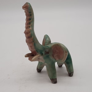 Könnte beinhalten: Eine Keramik-Elefantenfigur mit erhobenem Rüssel. Der Elefant ist überwiegend türkis mit rosa und braunen Akzenten an Ohren und Rüssel. Die Beine sind dunkelgrau. Die Figur ist ein dekorativer Gegenstand.