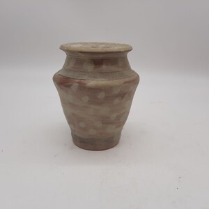 May include: A small, handcrafted ceramic vase with a speckled, earthy design. The vase features a light beige base with reddish-brown and white polka dot accents. The vase has a rounded body and a slightly flared rim.