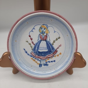 May include: A small, round, white ceramic plate with a blue and red painted design of a girl in a traditional dress. The plate has a pink rim and a blue and white floral pattern.