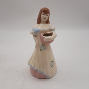 Puede incluir: Una figurita de cerámica de una mujer con un vestido blanco con detalles azules y rosas. Lleva una cesta rosa con un diseño floral. La figurita es de estilo vintage y sería una gran adición a cualquier decoración del hogar.