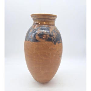 Puede incluir: Un jarrón de cerámica marrón con cuerpo redondeado y borde ligeramente ensanchado. La parte superior del jarrón presenta un patrón oscuro y moteado, mientras que la parte inferior es de color marrón claro liso. El jarrón tiene un aspecto rústico y artesanal.