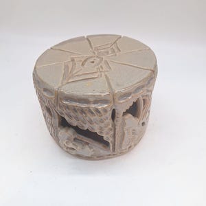 May include: A light gray ceramic candle holder with carved dragon and floral designs. The top has a geometric pattern with a central hole. The sides feature cut-out shapes, creating a decorative and functional piece.