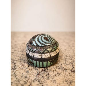 May include: A small, round, lidded ceramic container featuring a textured surface with a mix of turquoise, white, and dark brown glazes. The lid displays a swirling pattern, while the base is decorated with vertical stripes and a cross-hatched band.