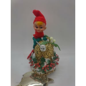 Puede incluir: Adorno de campana navideña vintage con un elfo con gorro y bufanda rojos. La campana es de purpurina dorada con acentos de acebo y bayas rojas. Un pájaro plateado está posado en la campana.