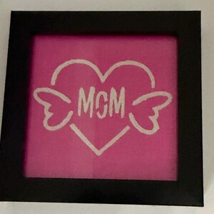 May include: A framed artwork with a pink background features a white heart with wings and the letters "MGM" inside. The frame is black. The artwork is a decorative piece.