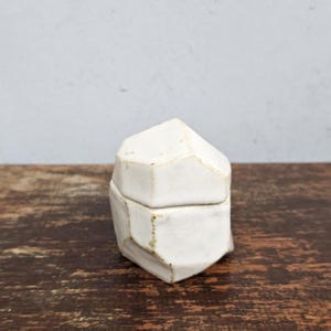 Peut inclure: Une boîte en céramique blanche avec un design géométrique à facettes. La boîte a un couvercle et une base, tous deux avec des bords irréguliers. La céramique a une finition mate et une couleur légèrement blanc cassé. La boîte est posée sur une surface en bois foncé.
