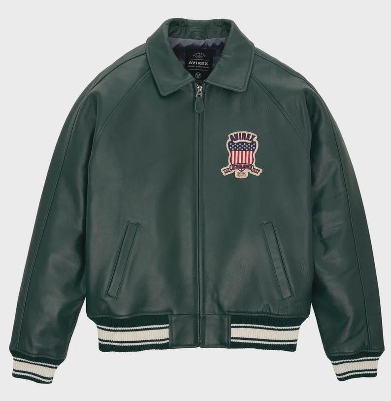 Puede incluir: Chaqueta de cuero verde oscuro con cremallera, cuello y pu&ntilde;os y dobladillo acanalados. La chaqueta presenta un parche en el pecho con el texto "AVIREX" y un dise&ntilde;o de bandera estadounidense. Dos bolsillos delanteros.