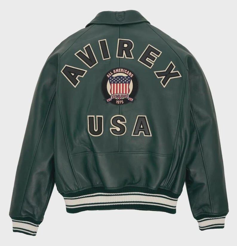 Puede incluir: Chaqueta de cuero verde oscuro con la palabra "AVIREX" arqueada en la parte superior de la espalda. Debajo, un parche con las palabras "ALL AMERICANS" y "ESTABLISHED 1975" rodeando una bandera estadounidense. La palabra "USA" est&aacute; debajo. La chaqueta tiene pu&ntilde;os y dobladillo a rayas blancas y negras.
