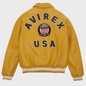 Puede incluir: Chaqueta de cuero amarillo mostaza con la palabra "AVIREX" arqueada en la espalda, sobre un parche circular con las palabras "ALL AMERICANS ESTABLISHED 1975" y un dise&ntilde;o de bandera estadounidense. La palabra "USA" est&aacute; debajo. Los pu&ntilde;os y el dobladillo tienen rayas blancas y crema.