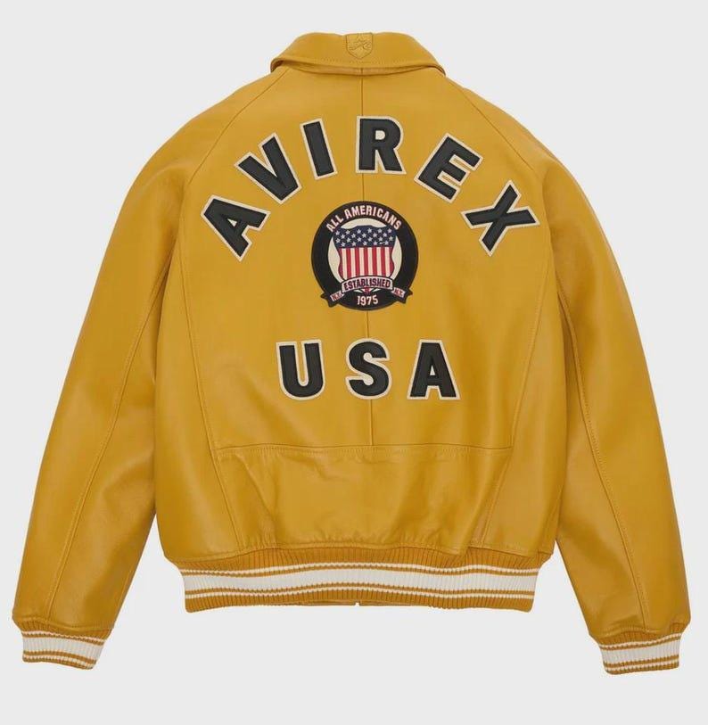Puede incluir: Chaqueta de cuero amarillo mostaza con la palabra "AVIREX" arqueada en la espalda, sobre un parche circular con las palabras "ALL AMERICANS ESTABLISHED 1975" y un dise&ntilde;o de bandera estadounidense. La palabra "USA" est&aacute; debajo. Los pu&ntilde;os y el dobladillo tienen rayas blancas y crema.