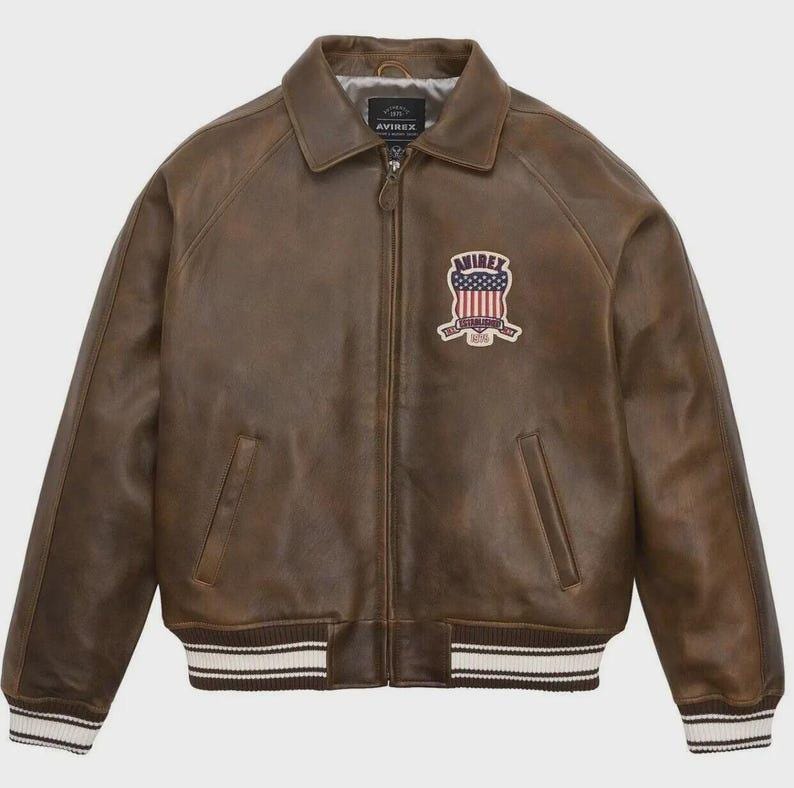 Puede incluir: Chaqueta de cuero marr&oacute;n con cremallera, cuello y pu&ntilde;os y dobladillo acanalados con rayas blancas. Cuenta con un parche en el pecho con el texto "AVIREX ESTABLISHED 1975" y un dise&ntilde;o de bandera estadounidense.