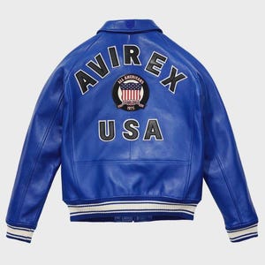 Puede incluir: Chaqueta de cuero azul vibrante con la palabra "AVIREX" en letras blancas en la espalda. Debajo, un parche circular muestra "ALL AMERICANS ESTABLISHED 1975" y un dise&ntilde;o de la bandera estadounidense. La palabra "USA" est&aacute; debajo.