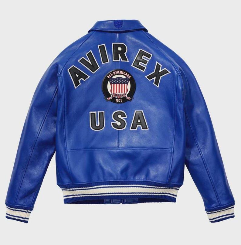 Puede incluir: Chaqueta de cuero azul vibrante con la palabra "AVIREX" en letras blancas en la espalda. Debajo, un parche circular muestra "ALL AMERICANS ESTABLISHED 1975" y un dise&ntilde;o de la bandera estadounidense. La palabra "USA" est&aacute; debajo.