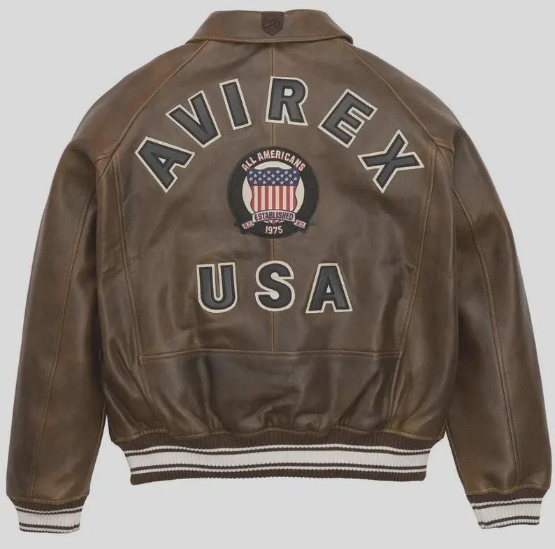 Puede incluir: Chaqueta de cuero marr&oacute;n con la palabra "AVIREX" en los hombros y "USA" en la parte inferior. Un parche circular en el centro presenta las palabras "ALL AMERICANS" y un dise&ntilde;o de bandera estadounidense. La chaqueta tiene un dobladillo a rayas blanco y marr&oacute;n.