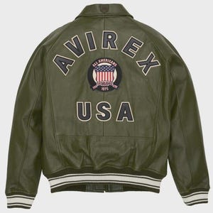Puede incluir: Chaqueta de cuero verde oliva con la palabra "AVIREX" arqueada en la espalda, encima de un parche circular con la bandera americana y las palabras "ALL AMERICANS ESTABLISHED 1975". La palabra "USA" est&aacute; debajo. La chaqueta tiene un dobladillo a rayas blanco y negro.