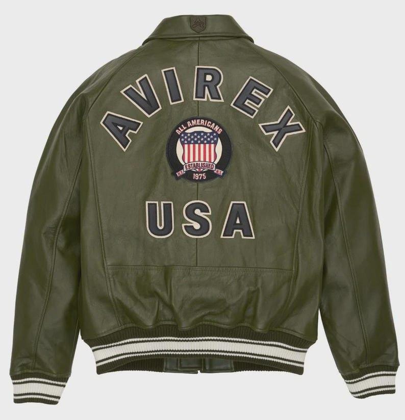 Puede incluir: Chaqueta de cuero verde oliva con la palabra "AVIREX" arqueada en la espalda, encima de un parche circular con la bandera americana y las palabras "ALL AMERICANS ESTABLISHED 1975". La palabra "USA" est&aacute; debajo. La chaqueta tiene un dobladillo a rayas blanco y negro.